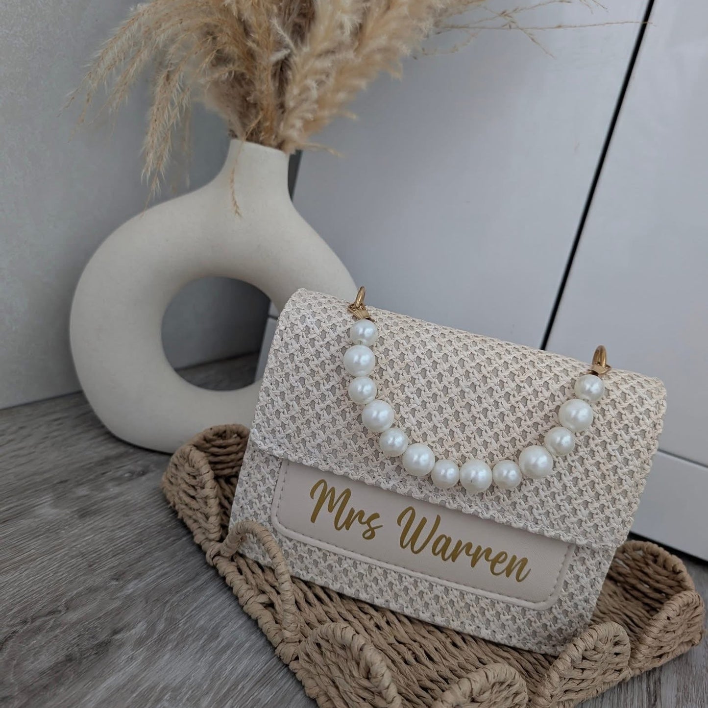 Beige handbag with pearl details and 'Mrs Warren' engraving on a woven mat with dried plants in the background.