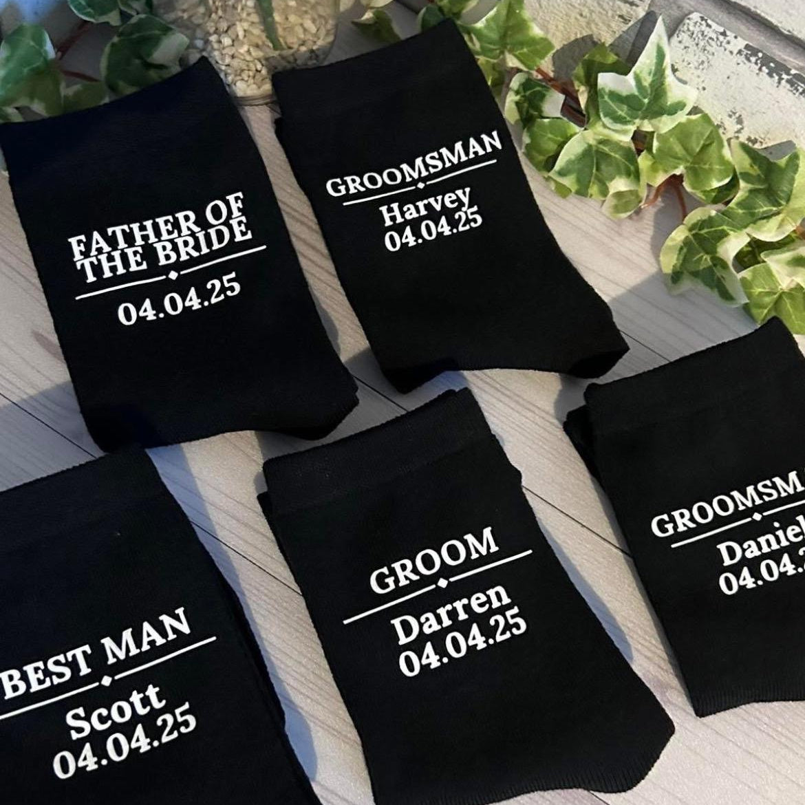 Black socks with personalized text for wedding party members on a wooden surface.