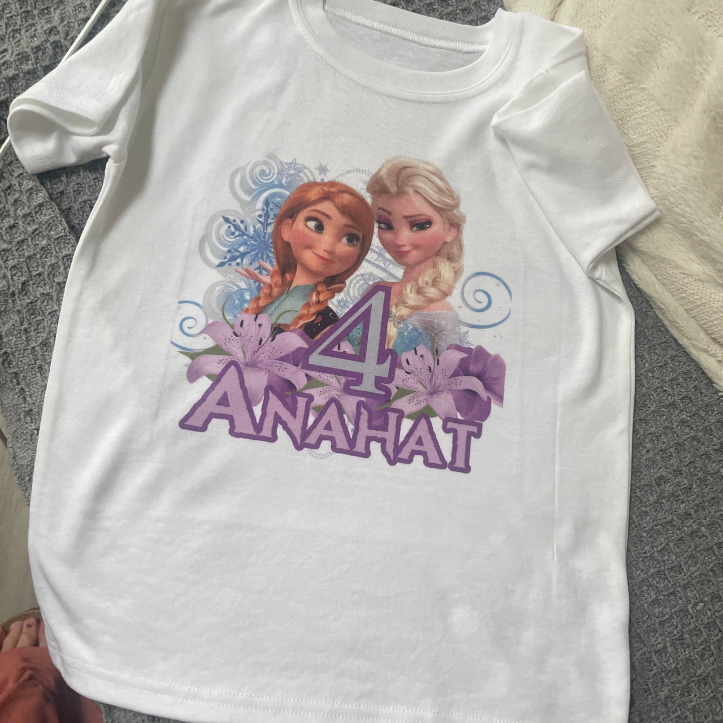 White t-shirt with cartoon characters and 'Anna & Elsa' text on a textured surface
