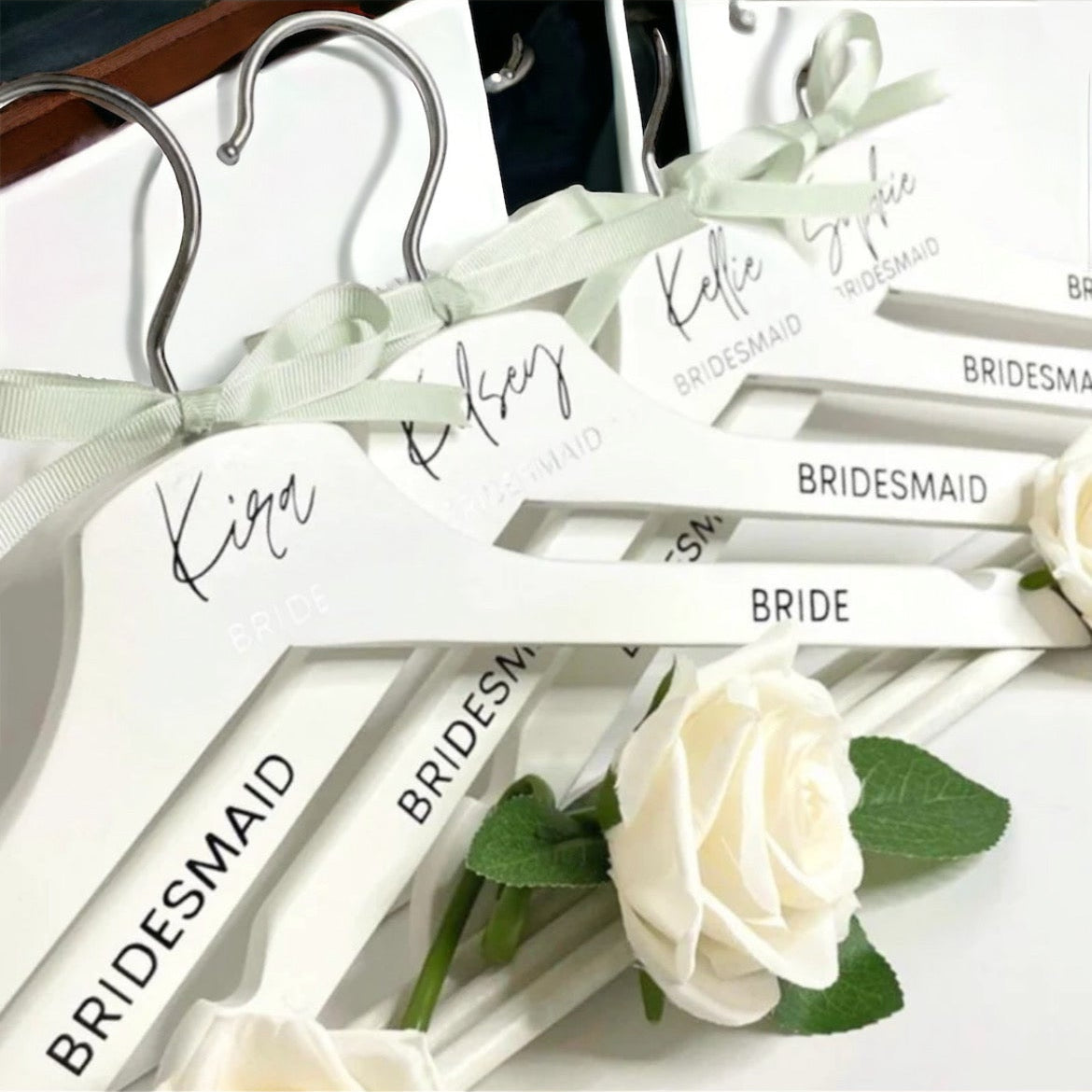 Hangers with 'Bride', 'Bridesmaid', and other labels on a white background with a white rose.