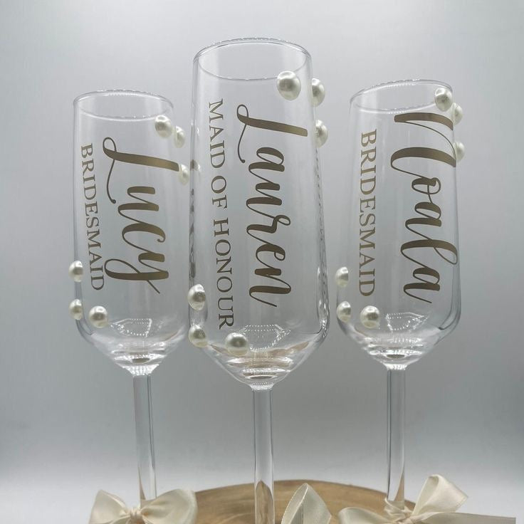 Three champagne flutes with names engraved on them, each tied with a bow, on a wooden stand.
