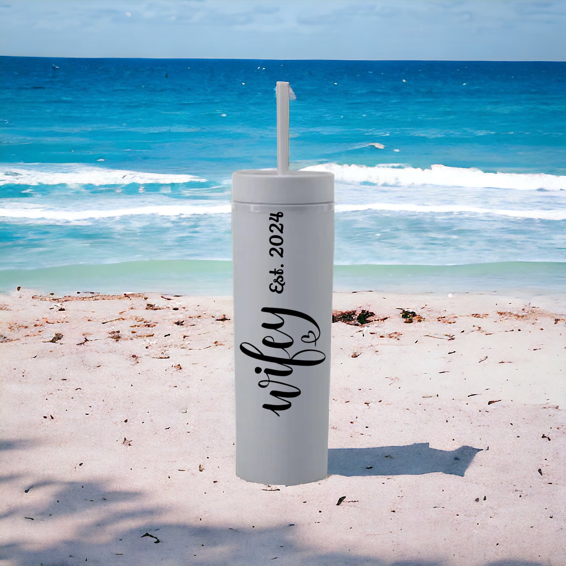 White tumbler with 'wifey' and 'Eck 2024' text on a beach with ocean in the background