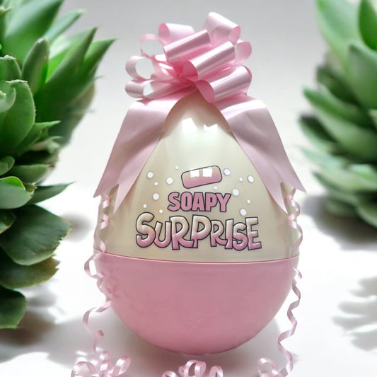 A pink Easter egg with the text 'SOAPY SURPRISE' on it, decorated with a pink bow and placed on a surface with green plants in the background.